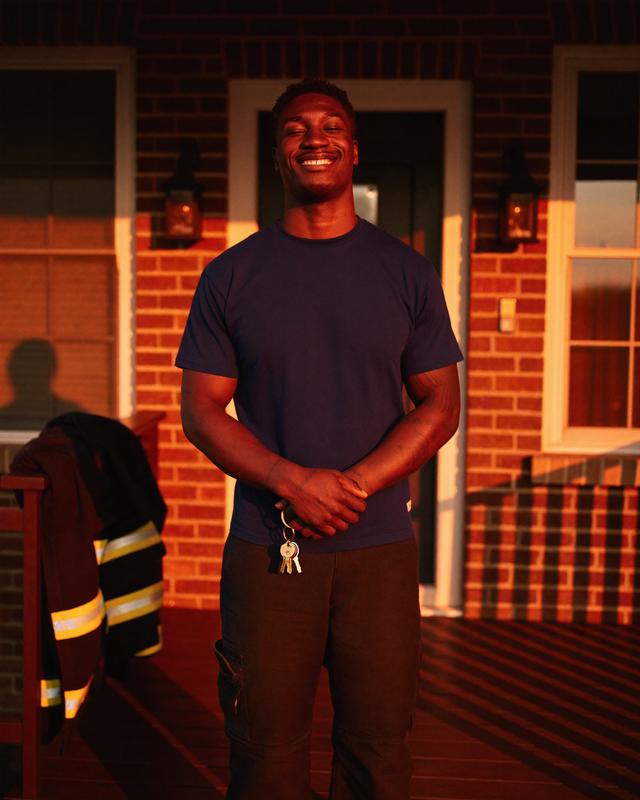 A firefighter in a plain navy tee holds house keys on an ember-lit porch, turnout coat over the rail.