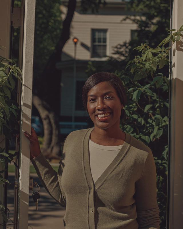 A teacher in a sage cardigan stands at her open green front door, keys hanging in the lock.